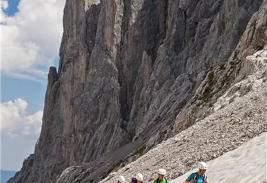 Eine Gruppe von Wanderern erklimmt einen felsigen Hang in den Bergen. Im Hintergrund sind majestätische Felsformationen und ein blauer Himmel zu sehen.
