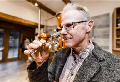 Ein Mann riecht an einem Glas mit klarer Flüssigkeit in einer modernen Destillerie. Im Hintergrund sind kupferne Geräte und Holzstrukturen zu sehen.