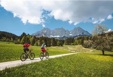 Zwei Radfahrer fahren auf einem Weg durch eine grüne Wiese. Im Hintergrund sind beeindruckende Berge und ein blauer Himmel zu sehen.