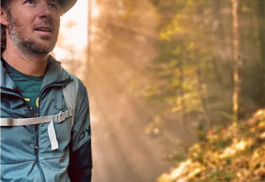 Ein Wanderer in einem Wald mit sanftem Licht, das durch die Bäume scheint. Er trägt eine Wanderausrüstung und einen Hut.