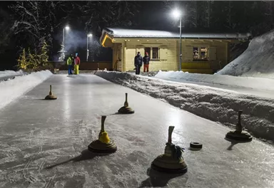 A snow track for curling at night, illuminated by lights. In the background, there are several people and a wooden cabin.