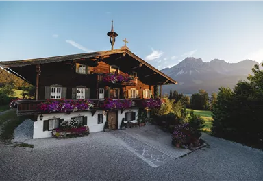 Ein traditionelles alpenländisches Haus mit schönen blühenden Blumen auf dem Balkon. Im Hintergrund sind beeindruckende Berge und ein klarer Himmel zu sehen.