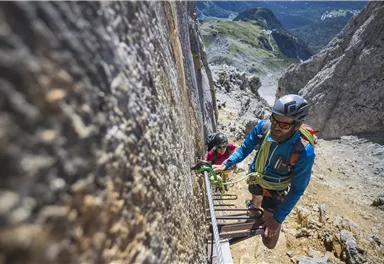 Zwei Bergsteiger erklimmen eine steile Felswand in einer beeindruckenden Berglandschaft. Die Sonne scheint und die Aussicht auf die Berge ist atemberaubend.