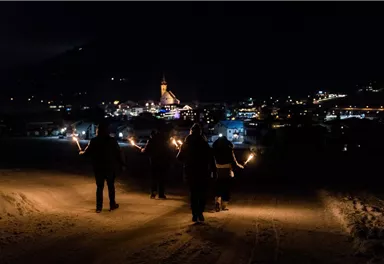 Eine Gruppe von Menschen geht mit Fackeln durch den Schnee in einer Stadt bei Nacht. Im Hintergrund sind beleuchtete Gebäude und eine Kirche zu sehen.