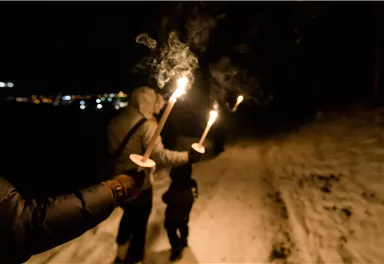 Eine Gruppe von Menschen geht nachts mit Fackeln auf einem verschneiten Weg. Die Atmosphäre wirkt festlich und geheimnisvoll.