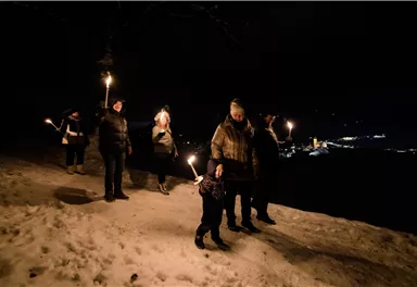 Eine Gruppe von Menschen geht nachts mit Fackeln durch eine schneebedeckte Landschaft. Im Hintergrund sind Lichter einer Stadt zu sehen.