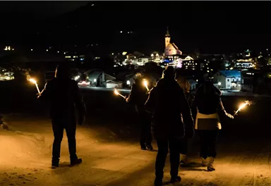 Eine Gruppe von Personen steht im Dunkeln und hält Fackeln. Im Hintergrund ist eine beleuchtete Stadt mit einer Kirche zu sehen.