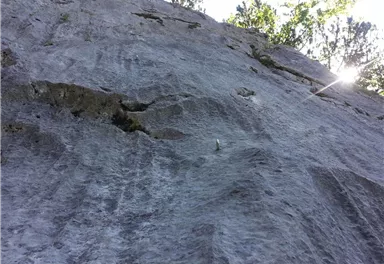 Eine steile Felswand mit einer strukturierten Oberfläche. Im Hintergrund sind helle Sonnenstrahlen und einige Bäume zu sehen.