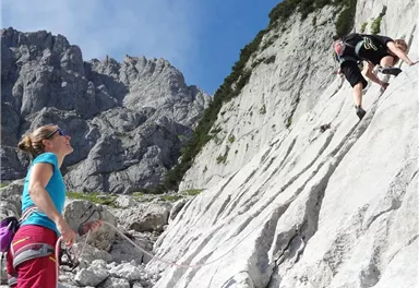 Eine Klettererin sichert einen Kletterer an einer steilen Felswand. Im Hintergrund sind beeindruckende Berggipfel zu sehen.