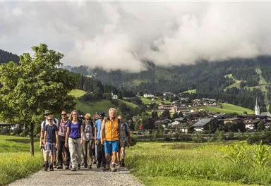 Eine Gruppe von Wanderern geht auf einem Weg durch eine grüne Landschaft. Im Hintergrund sind Berge und ein kleines Dorf mit Wolken am Himmel zu sehen.