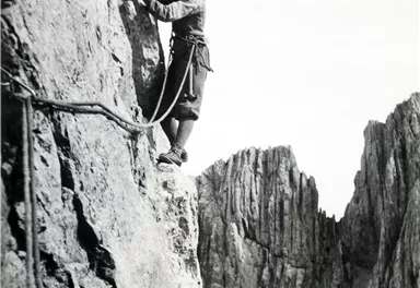Ein Bergsteiger klettert eine steile Felswand. Im Hintergrund sind beeindruckende Berggipfel zu sehen.
