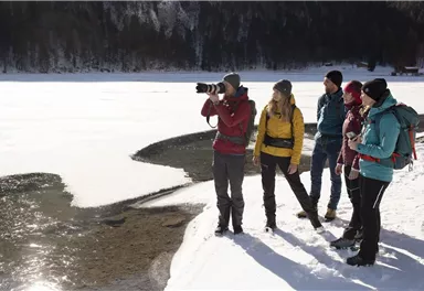 Eine Gruppe von fünf Personen steht am Ufer eines zugefrorenen Sees. Ein Mann fotografiert die Szene, während die anderen die Aussicht genießen.