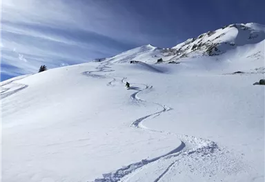 Eine verschneite Berglandschaft mit einem Skifahrer, der frische Spuren im Schnee hinterlässt. Im Hintergrund sind schneebedeckte Gipfel und ein blauer Himmel zu sehen.