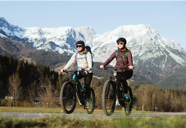 Zwei Radfahrerinnen fahren auf einem Weg inmitten einer malerischen Landschaft. Im Hintergrund sind schneebedeckte Berge und ein klarer Himmel zu sehen.