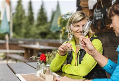 Zwei Frauen heben ihre Gläser in einer gemütlichen Außenumgebung. Im Hintergrund sind Bäume und eine rustikale Terrasse zu sehen.