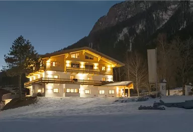 Ein gemütliches Chalet in den Bergen, beleuchtet in der Dämmerung. Es liegt in einer schneebedeckten Landschaft, umgeben von Bäumen und Bergen.