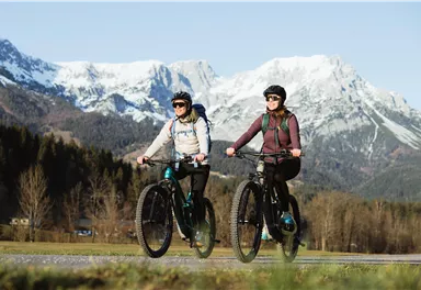 Zwei Personen fahren auf Mountainbikes in einer malerischen Berglandschaft. Im Hintergrund sind schneebedeckte Berge und eine grüne Wiese zu sehen.