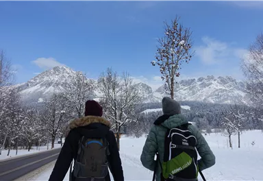 Zwei Wanderer stehen vor einer schneebedeckten Berglandschaft. Der Himmel ist klar und blau, die Umgebung wirkt friedlich und winterlich.