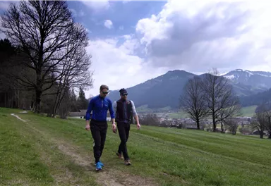 Zwei Personen gehen auf einem Wanderweg durch eine grüne Landschaft. Im Hintergrund sind Berge und eine bewölkte, aber helle Himmelsfarbe zu sehen.