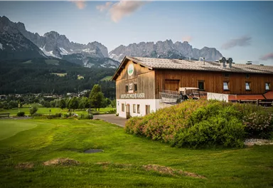 Ein rustikales Holzgebäude in einer grünen Umgebung, umgeben von Bergen. Die Szene zeigt eine friedliche Landschaft mit einem klaren Himmel.