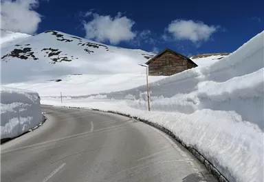 Eine schneebedeckte Straße mit hohen Schneewänden und einem Holzhaus in der Nähe. Der Himmel ist klar und blau mit einigen Wolken.