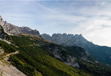 Eine beeindruckende Berglandschaft mit grünen Wäldern und hohen Gipfeln. Der Himmel ist blau mit leichten Wolken.
