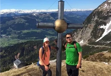 Zwei Wanderer posieren vor einem Gipfelkreuz in den Bergen. Im Hintergrund sind grüne Täler und schneebedeckte Berge zu sehen.