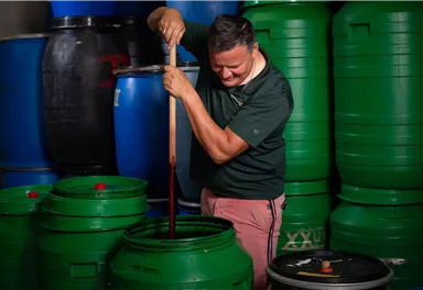 A man stirs with a long stick in a green barrel. In the background, several other green and blue barrels can be seen.