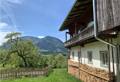 Ein traditionelles bayerisches Haus mit Holzstapel neben dem Garten. Im Hintergrund sind grüne Hügel und Berge unter einem klaren blauen Himmel zu sehen.