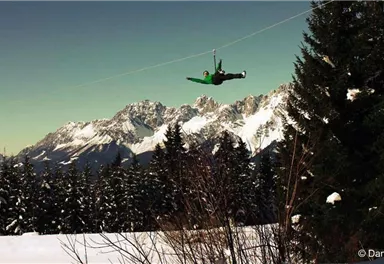 Ein Besucher schwebt an einer Seilrutsche über eine schneebedeckte Landschaft. Im Hintergrund sind majestätische Berge und Tannenbäume zu sehen.
