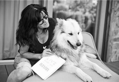 Eine Frau sitzt neben einem Hund und lächelt. Sie hält ein Notizbuch in der Hand und scheint zu schreiben.