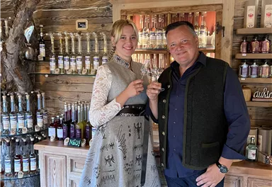 Ein Paar steht in einer gemütlichen Bar mit Holzregalen und verschiedenen Getränken. Beide halten ein Glas in der Hand und lächeln freundlich.