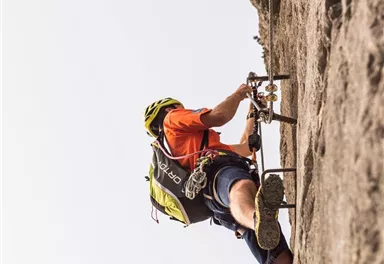 Ein Kletterer trägt einen Helm und versucht, eine steile Felswand zu erklimmen. Er ist in sportlicher Kleidung und befindet sich in einer herausfordernden Position.
