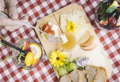 Ein rustikales Brett mit Käse, frischen Früchten und bunten Blumen. Im Hintergrund sind Getränke in Gläsern sichtbar, alles auf einem karierten Tischcloth.