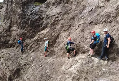 Eine Gruppe von Bergsteigern klettert an einer steilen Felswand. Sie tragen Helme und spezielle Ausrüstung für die Besteigung.
