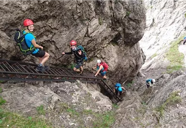 Eine Gruppe von Menschen klettert eine steile Felswand hinauf. Sie tragen Helme und Kletterausrüstung und sind von beeindruckenden Felsen umgeben.