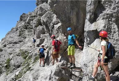 Eine Gruppe von Kletterern navigiert auf einem schmalen Pfad entlang einer felsigen Wand. Die Umgebung besteht aus beeindruckenden Felsen und einer klaren blauen Himmel.