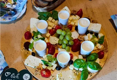 Ein eleganter Käseplatten mit verschiedenen Käsesorten, Trauben, Erdbeeren und Basilikum. In der Mitte sind kleine Becher platziert, die eine köstliche Soße enthalten.