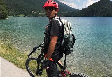 Ein Radfahrer steht am Ufer eines klaren Sees, umgeben von Bergen. Er trägt einen roten Helm und hat einen Rucksack auf dem Rücken.