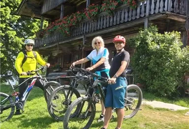 Drei Radfahrer stehen mit ihren Fahrrädern auf einer grünen Wiese vor einem holzverkleideten Haus. Die Sonne scheint und es ist ein klarer, blauer Himmel zu sehen.
