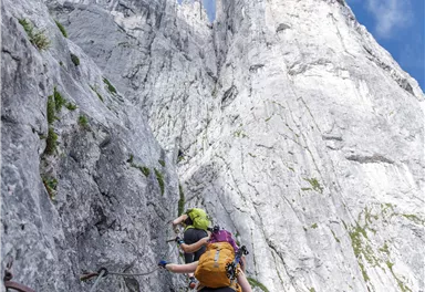 Zwei Kletterer steigen einen steilen, felsigen Hang hinauf. Die imposanten Felsformationen unter einem klaren blauen Himmel sind zu sehen.