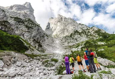 Eine Gruppe von Wanderern steht vor beeindruckenden Bergformationen. Die Landschaft ist grün und felsig, mit Wolken am Himmel.