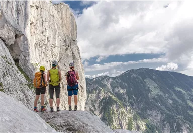 Drei Wanderer stehen auf einem steilen Felsen und blicken auf eine beeindruckende Berglandschaft. Der Himmel ist teils bewölkt und die Felsen sind hell und beeindruckend.