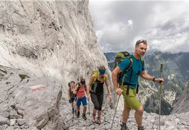 Eine Gruppe von Wanderern erklimmt einen steinigen Pfad in den Bergen. Die Umgebung ist felsig mit vielen hohen Felsen und einer beeindruckenden Aussicht.