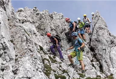 Eine Gruppe von Kletterern erklimmt einen felsigen Berg. Die Szene zeigt eine Mischung aus Felsen und Grün, umgeben von einem klaren Himmel.