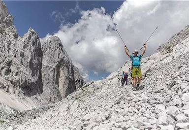 Zwei Wanderer auf einem steinigen Weg in den Bergen. Der vordere Wanderer feiert mit erhobenen Armen unter einem wolkigen Himmel.