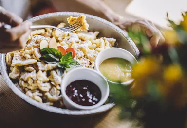 Ein Teller mit köstlichem Kaiserschmarrn, garniert mit Puderzucker und frischer Minze. Dazu gibt es zwei Schalen mit Apfelmus und Marmelade.