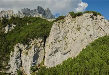 Eine beeindruckende Felslandschaft mit grünen Bäumen und steilen Gipfeln im Hintergrund. Der Himmel ist blau mit einigen Wolken.