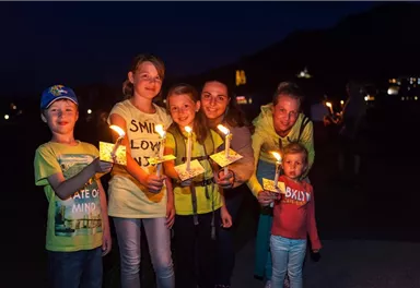 Eine Gruppe von Kindern und Erwachsenen hält Kerzen in der Dunkelheit. Sie lächeln und scheinen Freude an dem besonderen Moment zu haben.