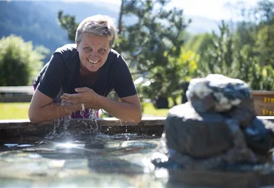 Eine lächelnde Frau steht an einem Brunnen mit klaren Wasser in einer grünen Umgebung. Im Hintergrund sind Bäume und Berge zu sehen.